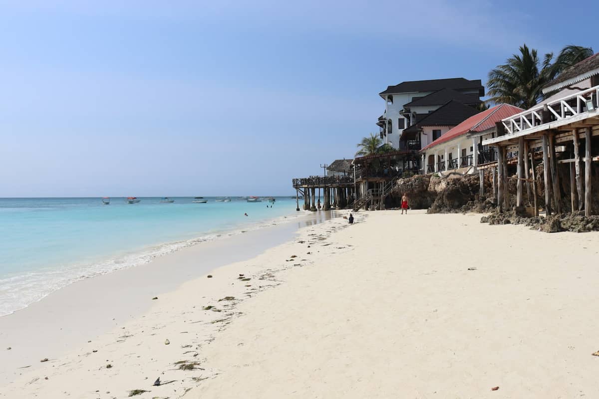 Pantai berpasir yang luas dengan air berwarna toska, restoran tepi pantai berpondasi tiang, dan beberapa orang berjalan di kejauhan di bawah langit biru.