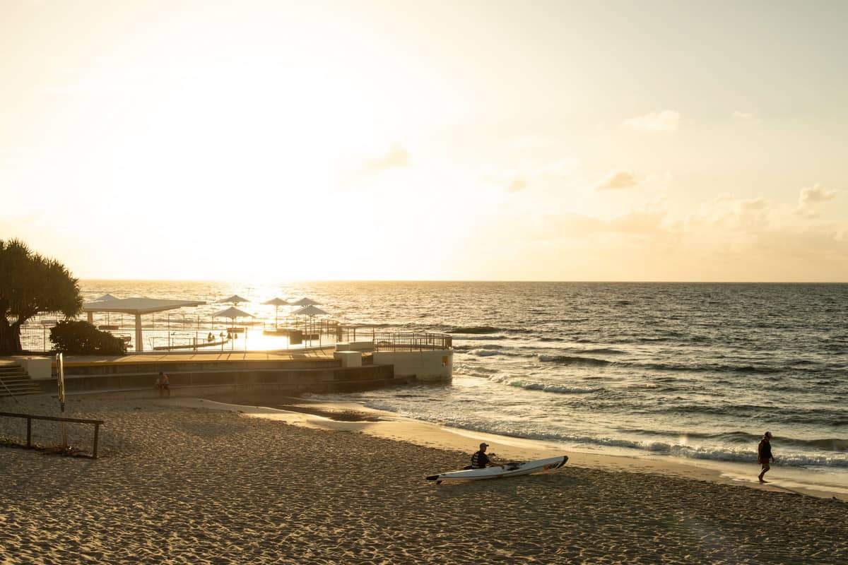 Sandy beach at sunset with a person launching a kayak and beach umbrellas near the water, with gentle surf and a relaxed atmosphere.