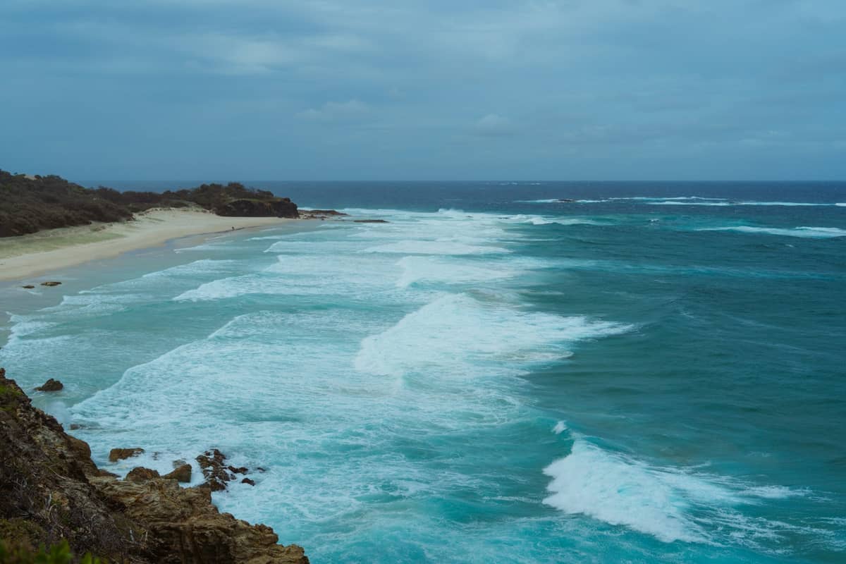 Wide sandy beach with turquoise waves and rocky headland under a cloudy sky, typical of North Stradbroke Island.