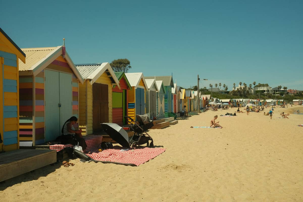 Una larga hilera de coloridas casetas de playa en Brighton sobre arena dorada, con visitantes disfrutando de un día soleado cerca de Melbourne.