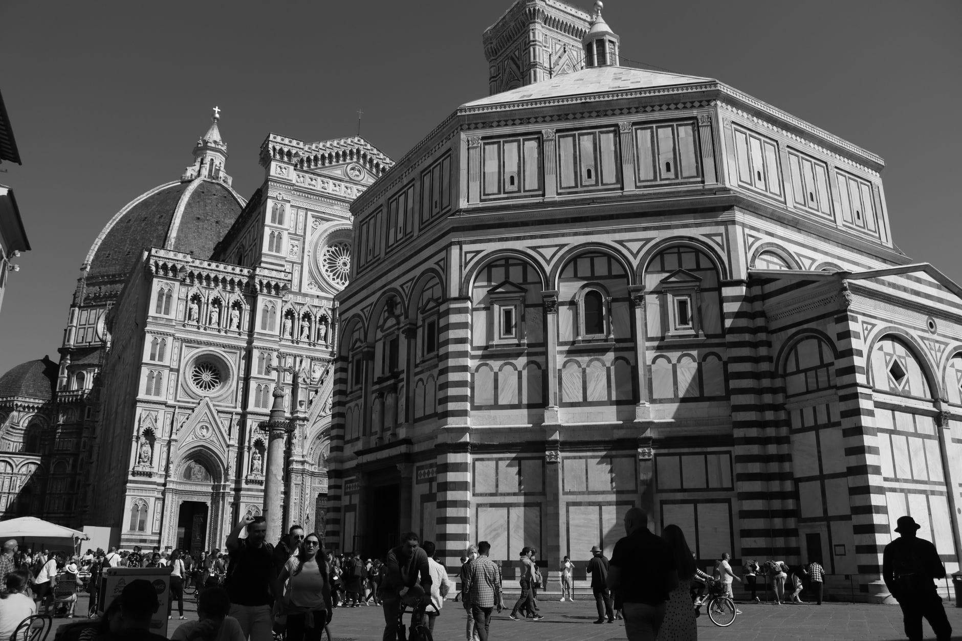 ภาพขาวดำของ Duomo, Baptistery และหอระฆังของเมือง Florence ที่มองเห็นจาก Piazza del Duomo โดยมีผู้คนเดินอยู่เบื้องหน้า