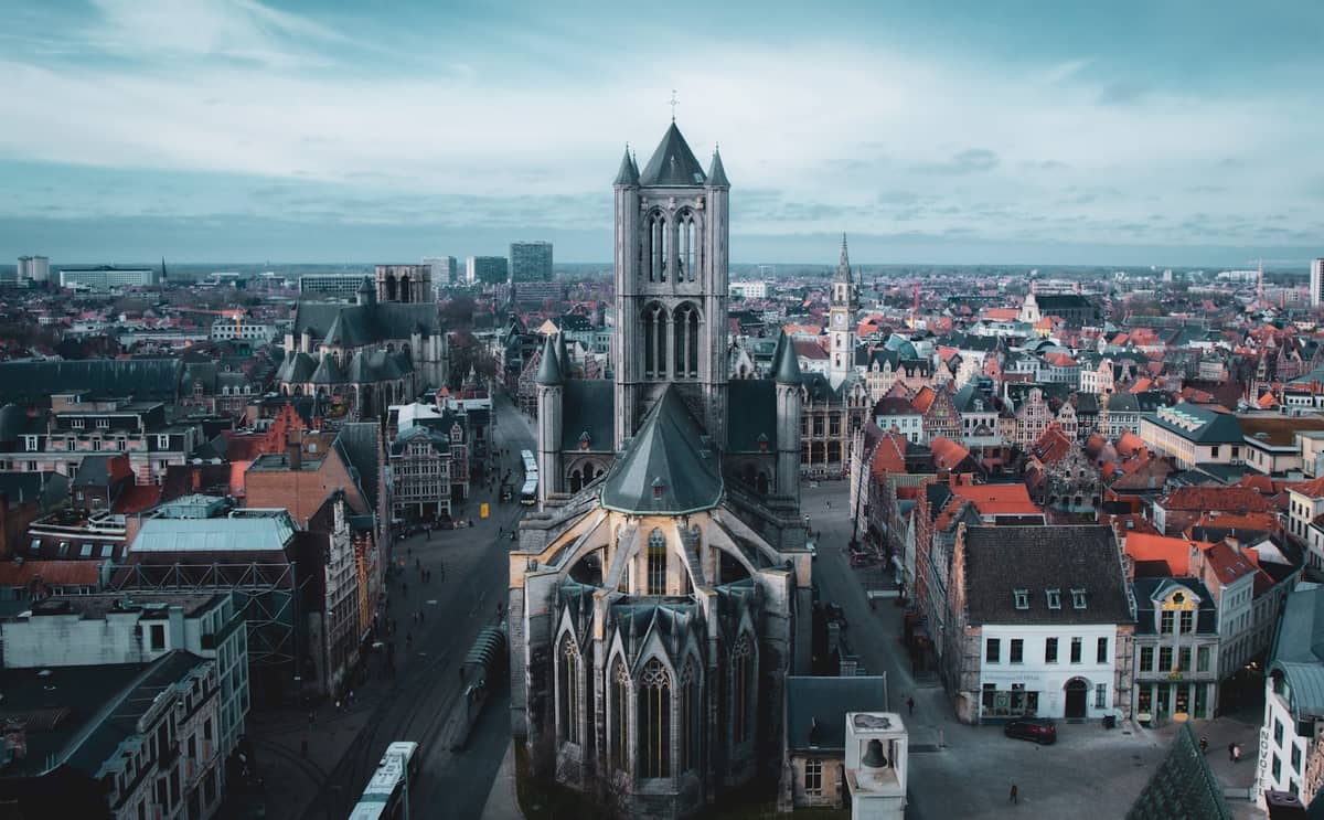Aerial view of St. Nicholas' Church in Ghent, Belgium, with its iconic towers dominating the cityscape under a bright blue sky.