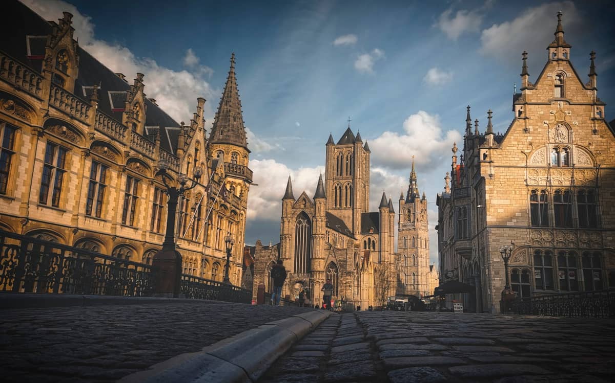 Iconic view of Ghent showing St. Nicholas' Church flanked by civic guild houses under a partly cloudy sky, with cobbled streets in the foreground.
