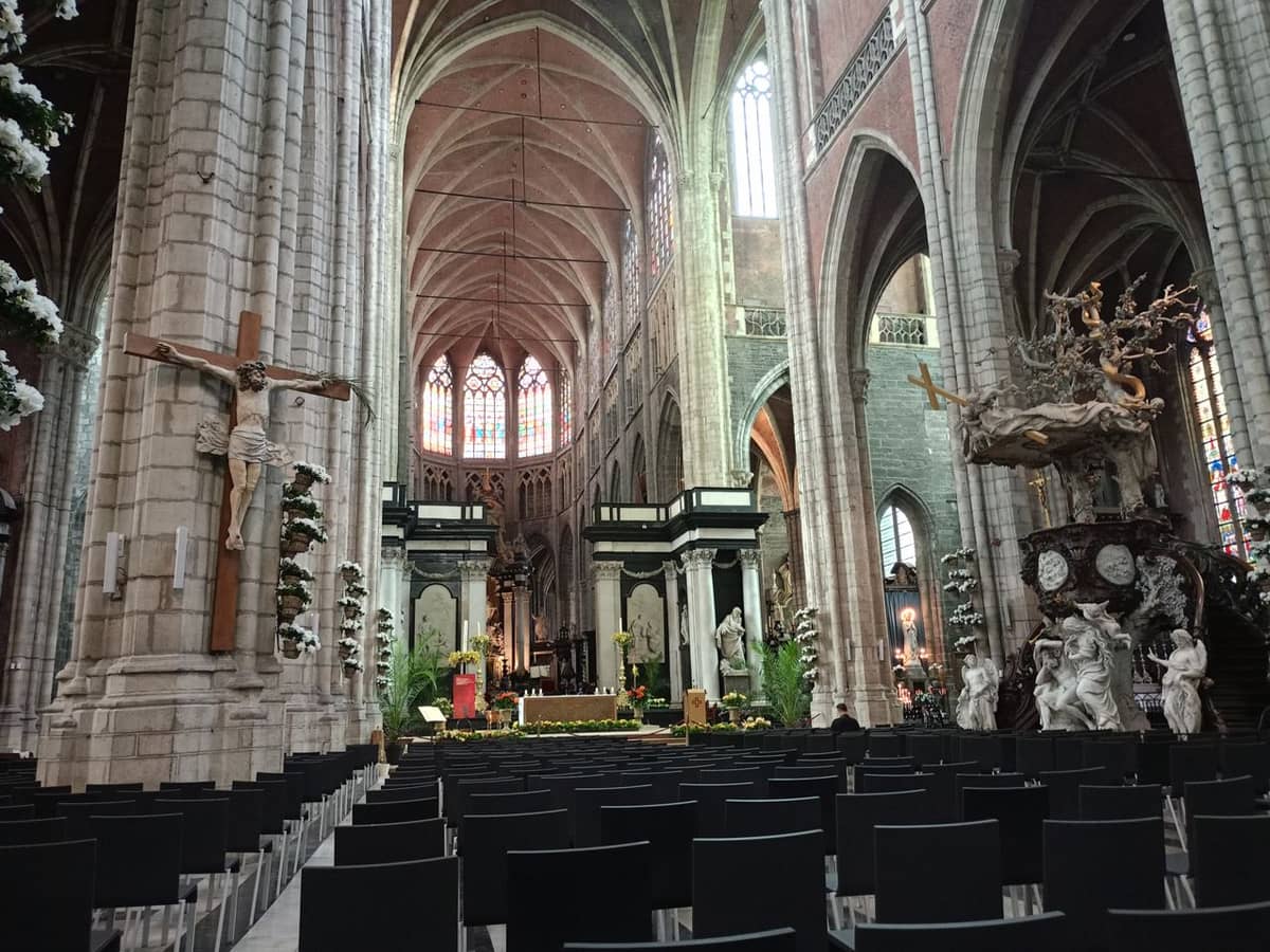 Interior of St. Bavo's Cathedral in Ghent, featuring rows of chairs, a large crucifix, ornate pulpit, and stained glass windows in a spacious gothic nave.