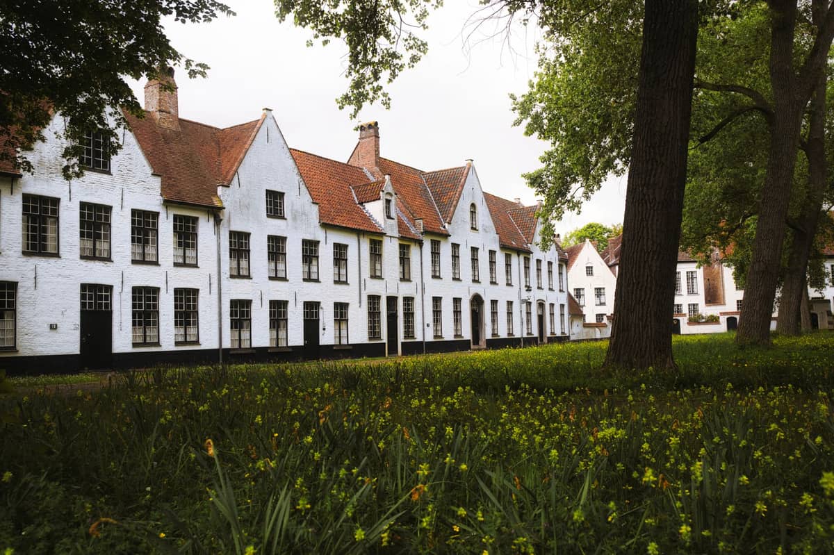 Long row of historic whitewashed houses with red tiled roofs, set behind a grassy area and trees, showing peaceful religious community architecture.