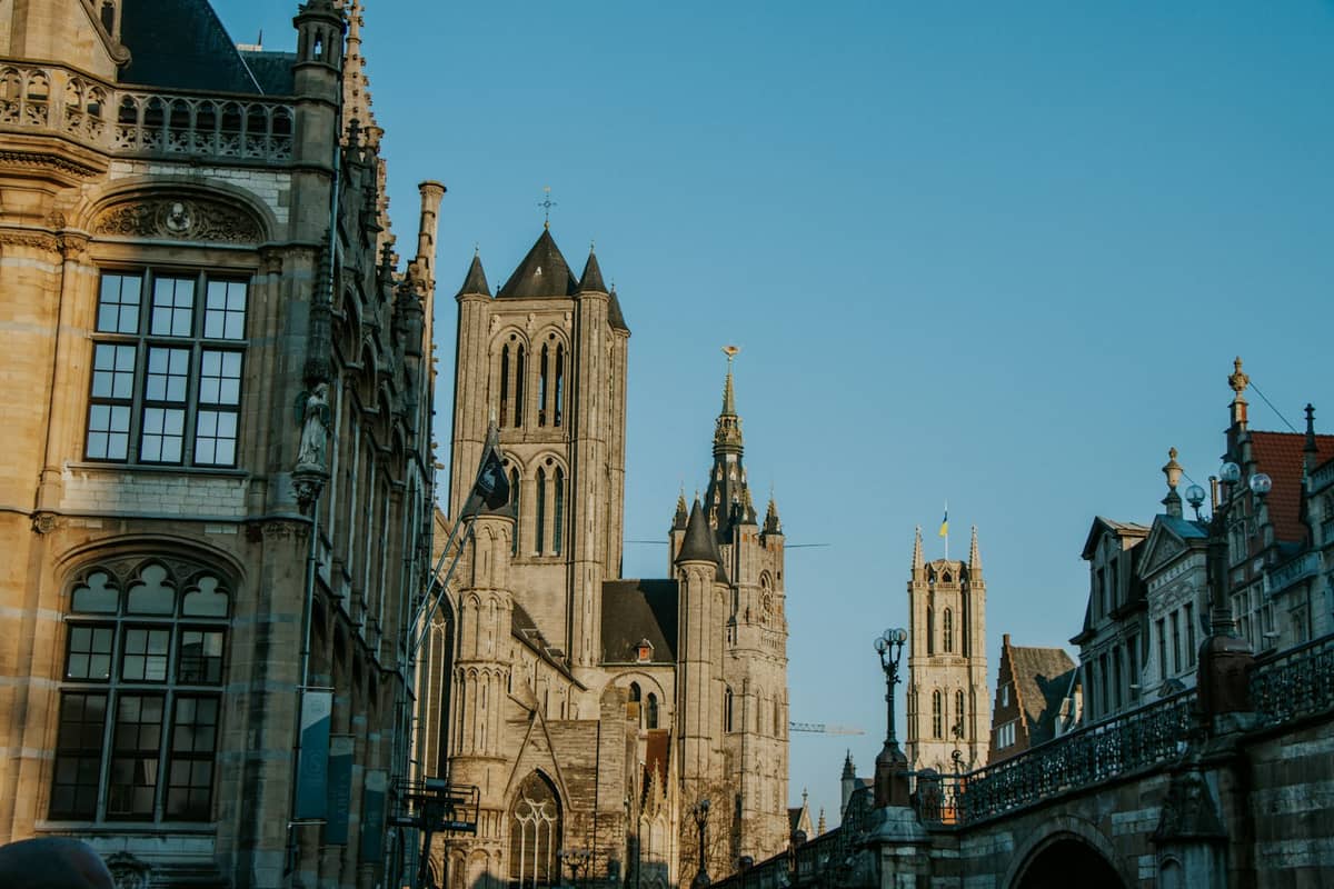 View from St Michael's Bridge showing Ghent's three towers: St Nicholas' Church, the Belfry, and St Bavo's Cathedral under a clear blue sky.