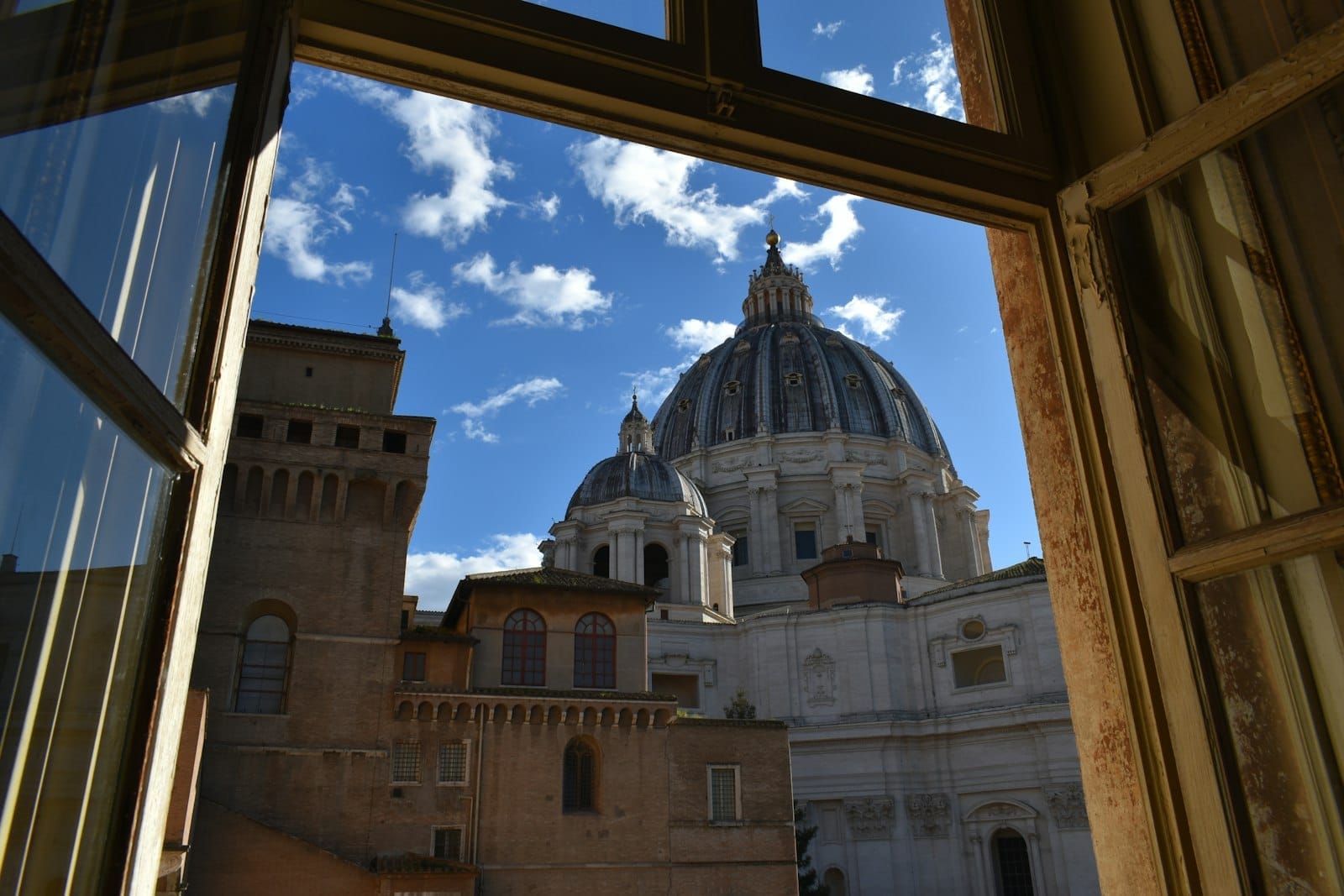 View of St. Peter's Basilica dome through an open window, with blue sky and clouds above Rome's rooftops.