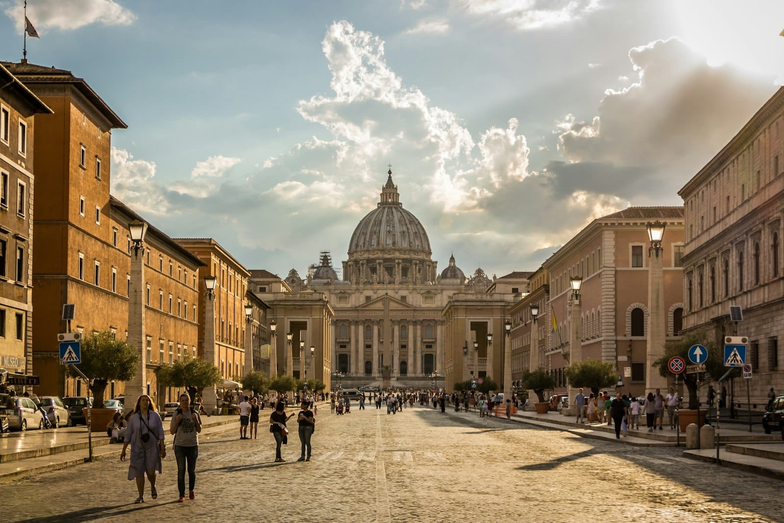 Wide view of St. Peter's Basilica with its iconic dome at the end of a bustling street in Rome, people walking toward the landmark.