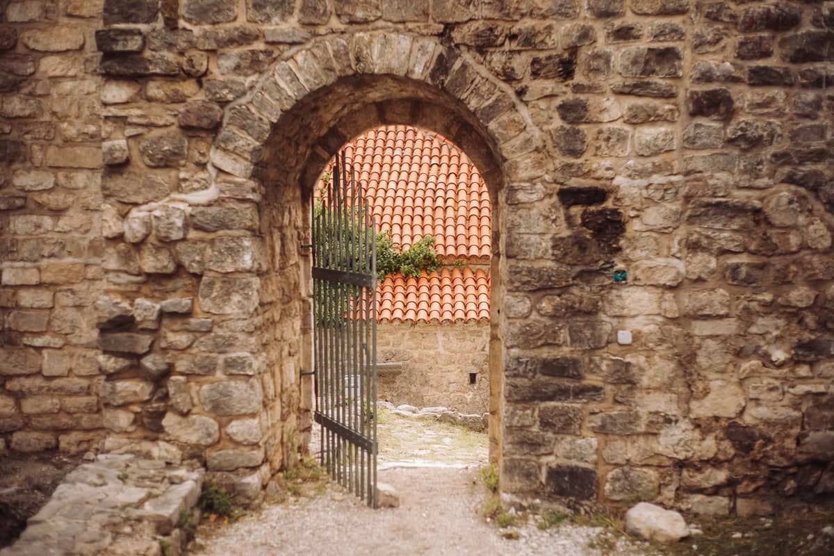 Muro de piedra medieval con una puerta en arco y rejas de hierro forjado, con un camino adoquinado y tejados de tejas rojas al fondo.