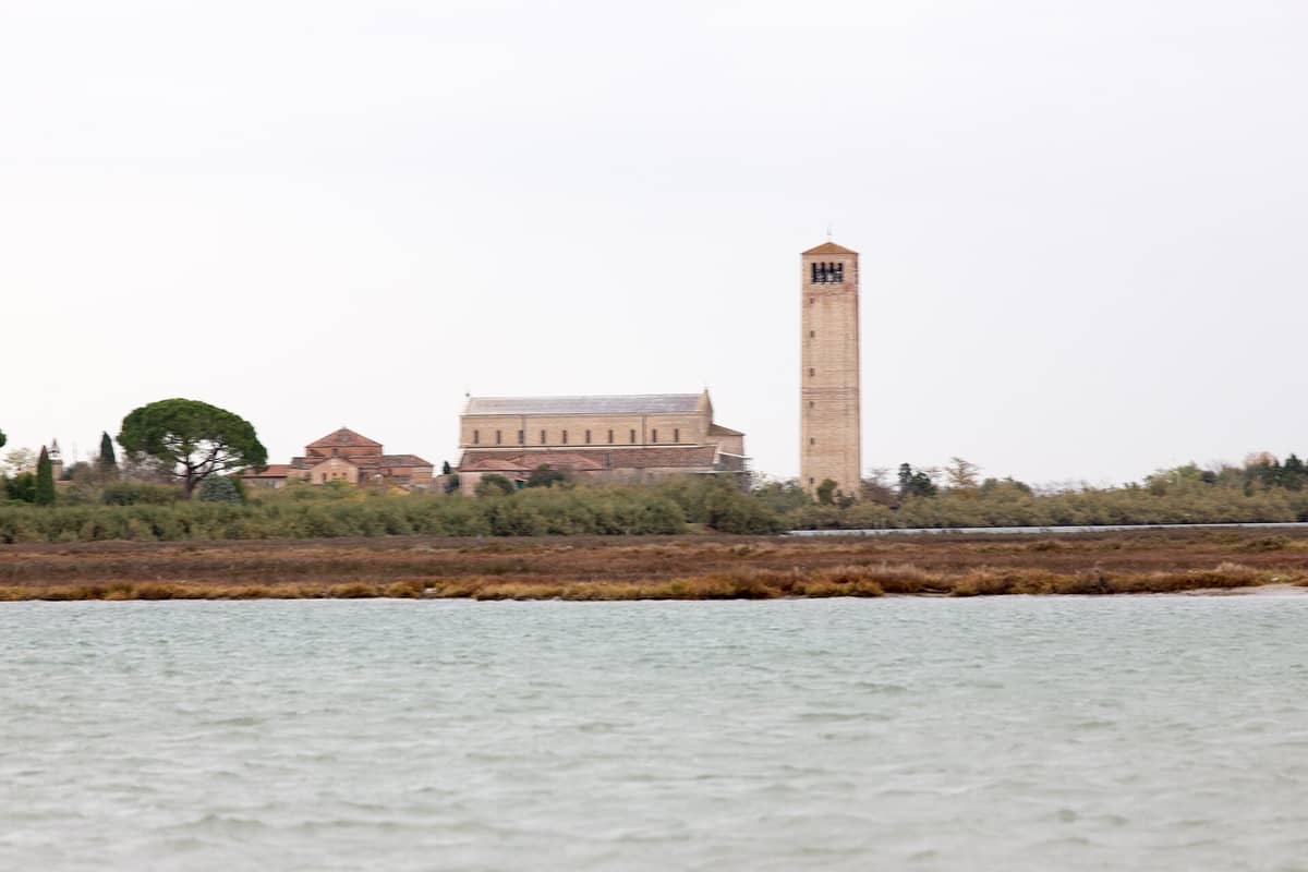 Blick über die Lagune zur antiken Basilica di Santa Maria Assunta und dem hohen Campanile auf der Insel Torcello, umgeben von Grün.
