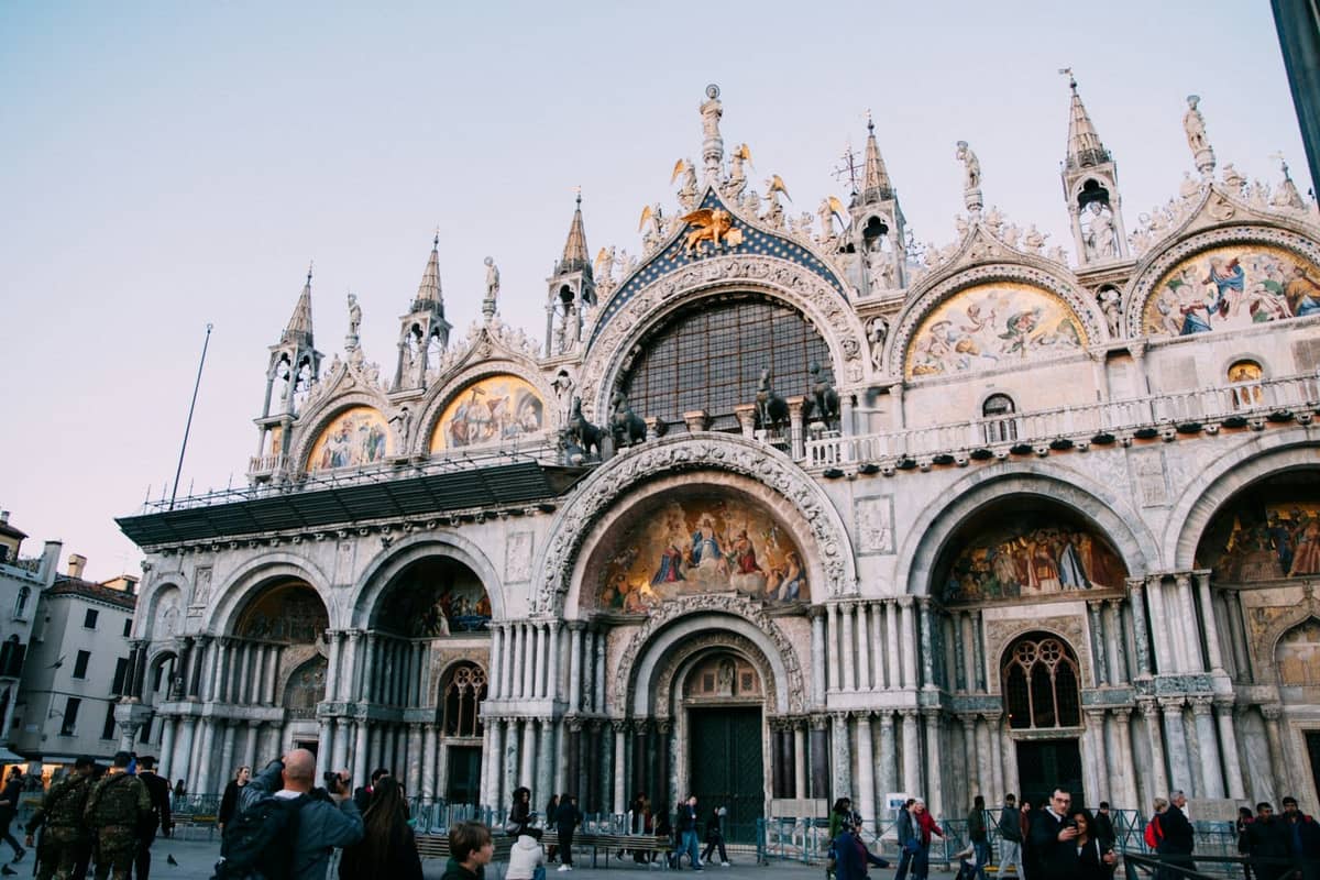 Fassade des Markusdoms in Venedig mit seinen reich verzierten Goldmosaiken und Kuppeln, vom Platz aus gesehen mit Menschen davor.