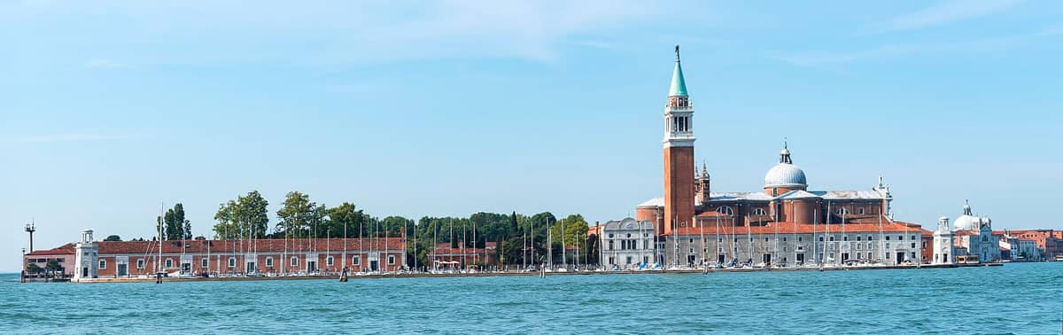 Weiter Blick auf die Kirche San Giorgio Maggiore auf ihrer eigenen Insel, über das Wasser unter blauem Himmel gesehen, Boote am Ufer vertäut.
