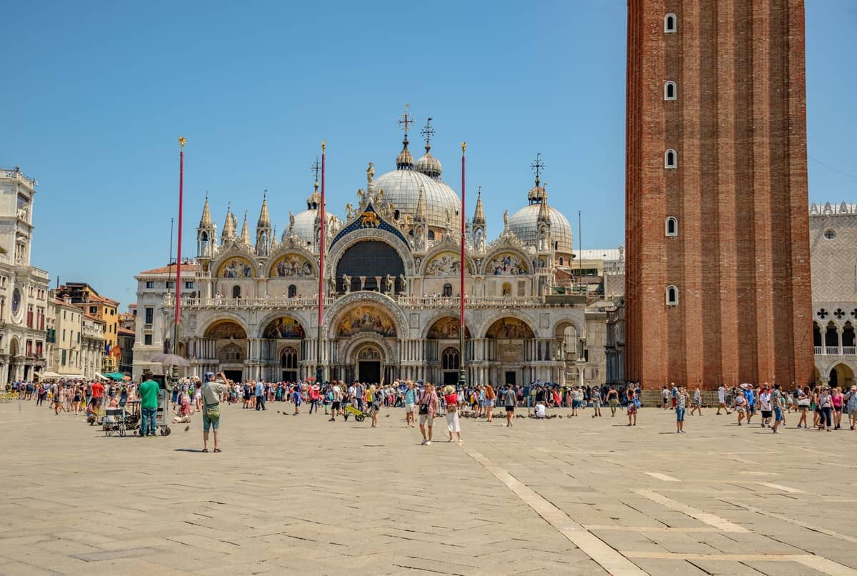 Weitblick auf die Markusbasilika in Venedig mit ihren reich verzierten Kuppeln und der Fassade, belebten Menschenmengen und einem Teil des Campanile bei hellem Tageslicht.
