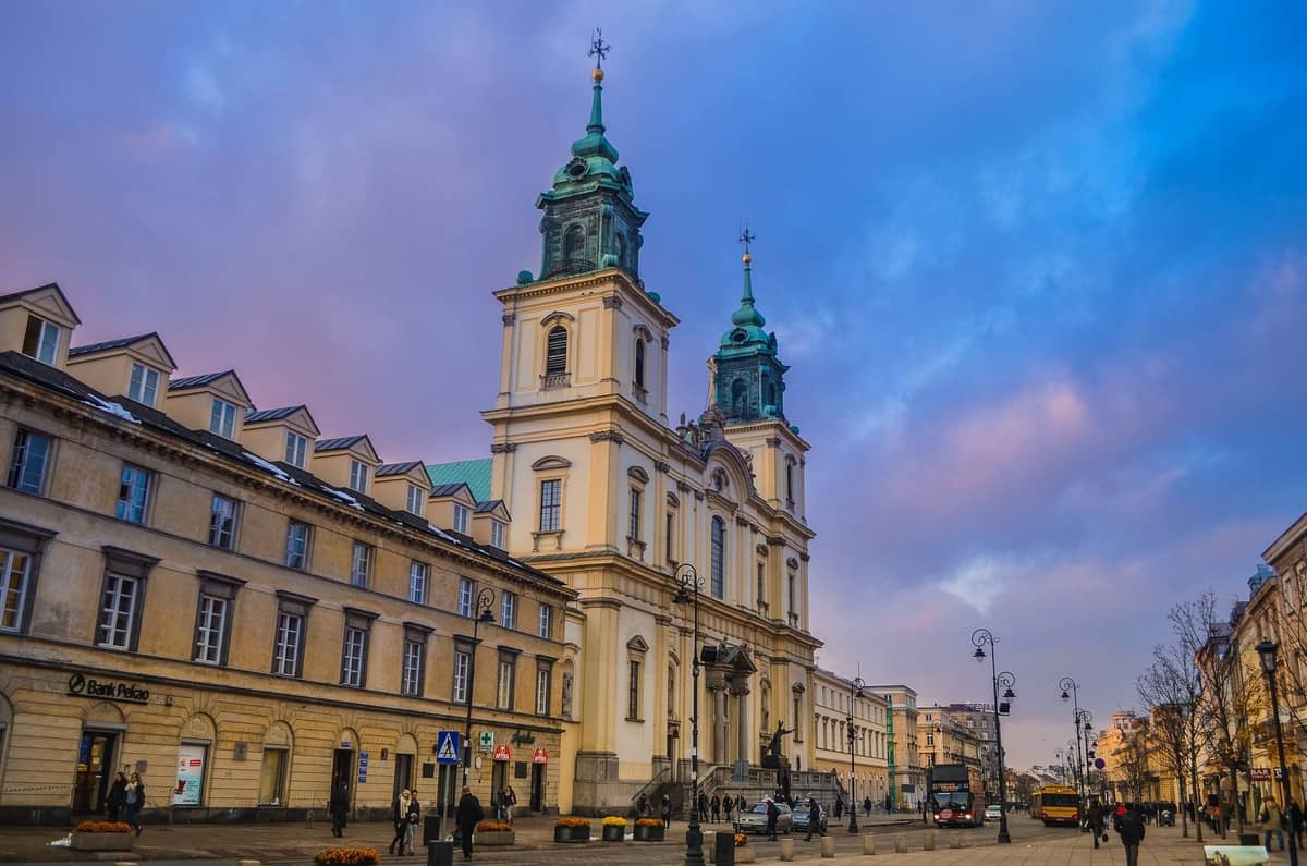 Kościół Świętego Krzyża z dwiema wieżami o zachodzie słońca przy Krakowskim Przedmieściu, tętniący życiem z ludźmi i klasycznymi latarniami ulicznymi w Warszawie.