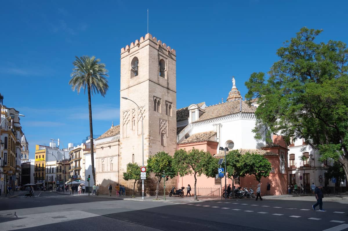 โบสถ์ประวัติศาสตร์ในเมือง Seville พร้อมหอระฆังสี่เหลี่ยมสไตล์ Mudéjar-Gothic ต้นปาล์ม หลังคาทาสีขาวและหลังคากระเบื้อง รวมถึงผู้คนที่เดินเล่นอยู่ใกล้เคียง