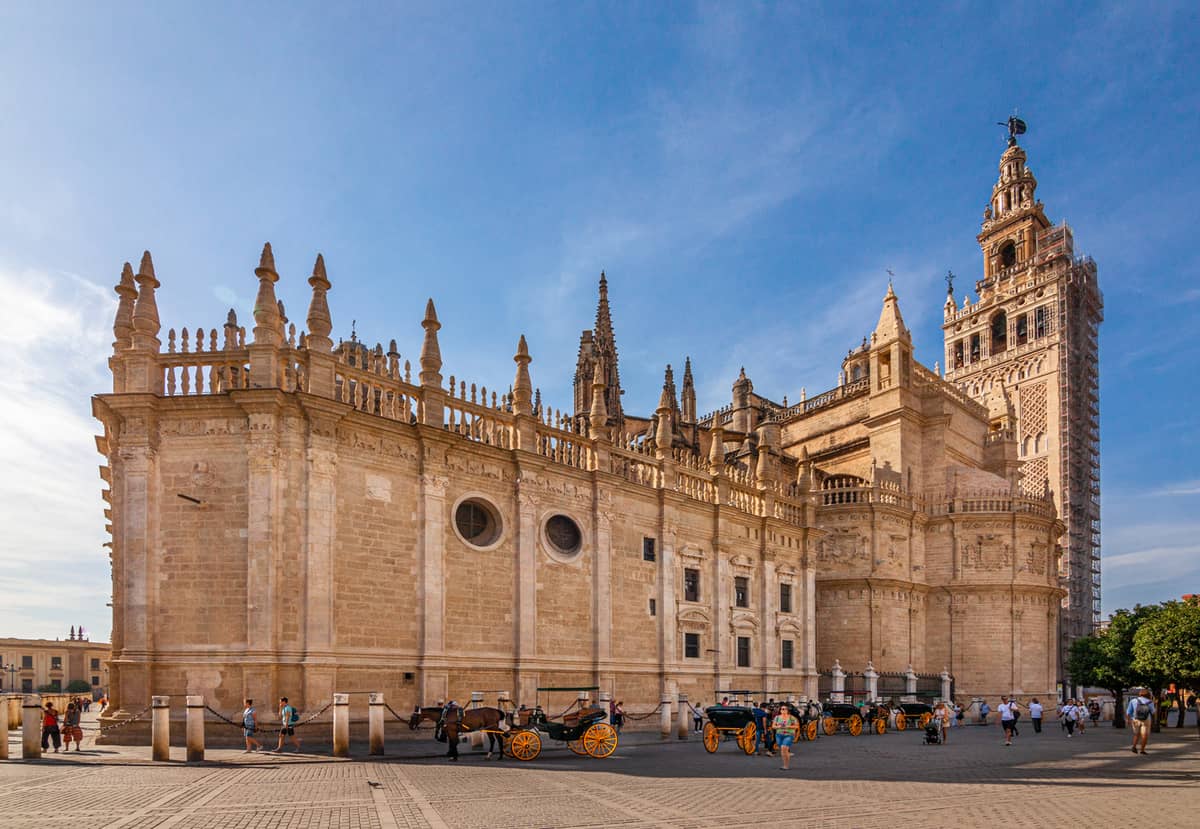 วิวกว้างของด้านนอกมหาวิหาร Seville พร้อมหอคอย Giralda ผนังหินและผู้คนเดินอยู่ในลานกว้างในวันที่แดดสดใส