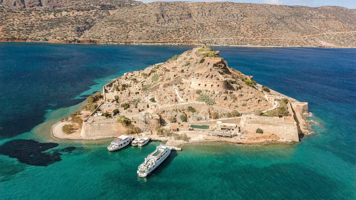Pemandangan udara benteng Pulau Spinalonga di lepas pantai dengan laut biru toska dan perahu wisata yang bersandar, lanskap pegunungan di latar belakang.