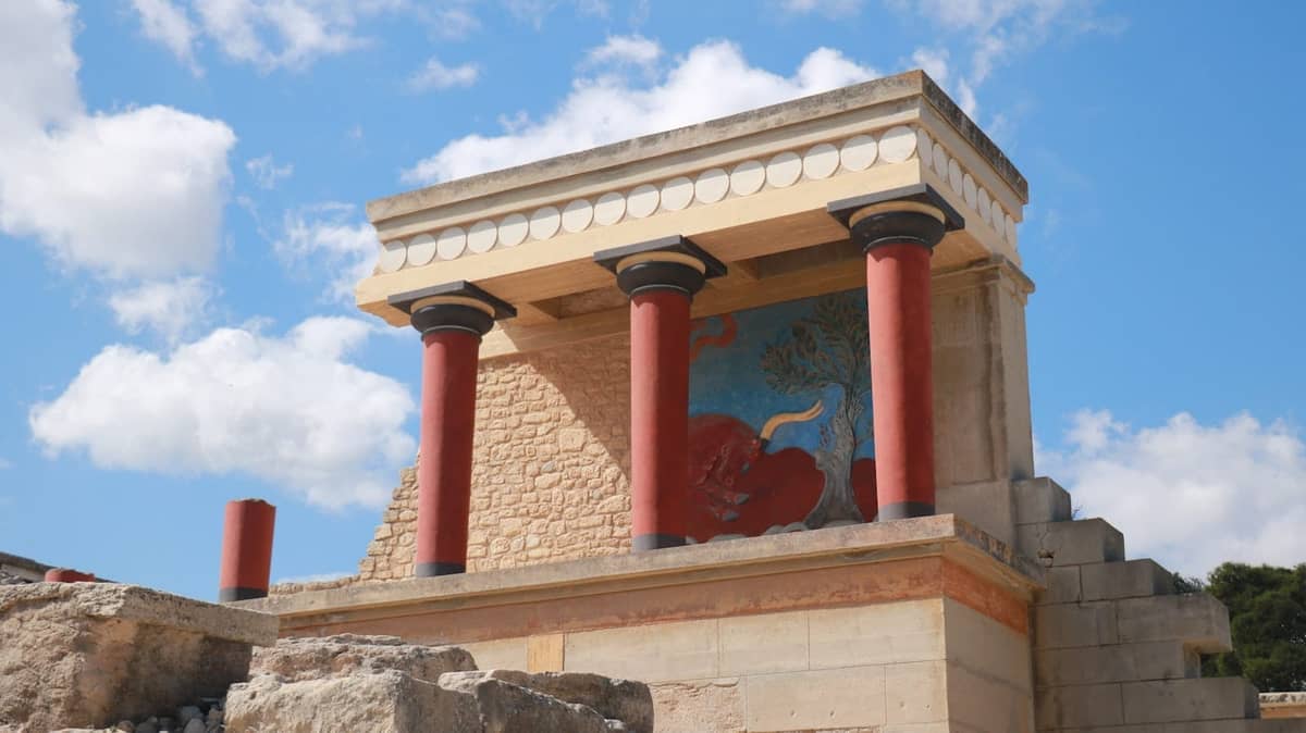 Pintu masuk utara yang direkonstruksi dari Istana Knossos, menampilkan kolom-kolom merah dan sebuah fresko, di bawah langit yang sebagian berawan.