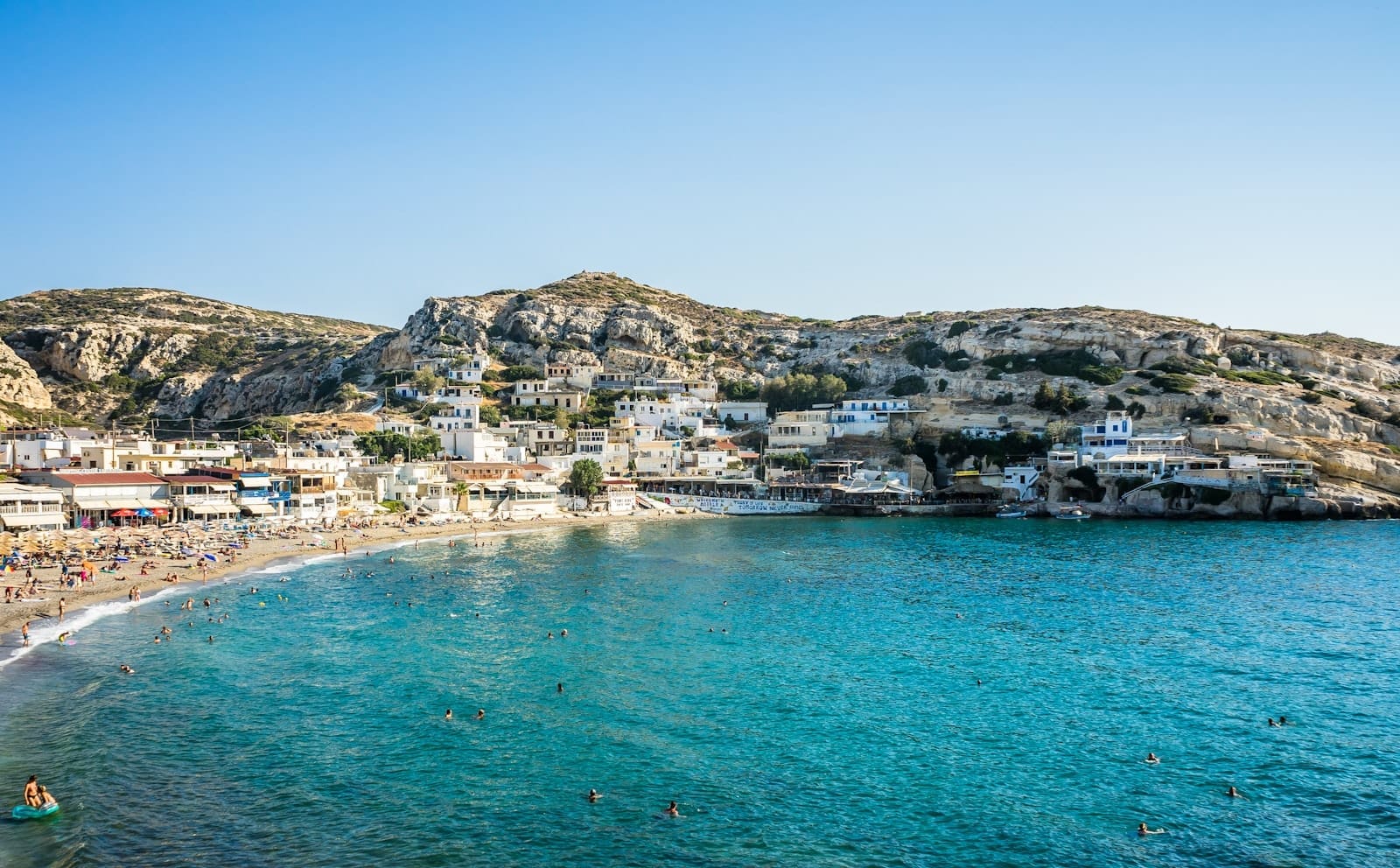 Vila costeira ensolarada em Creta com águas turquesa, banhistas, praia de areia, casas brancas e colinas rochosas sob um céu azul límpido.