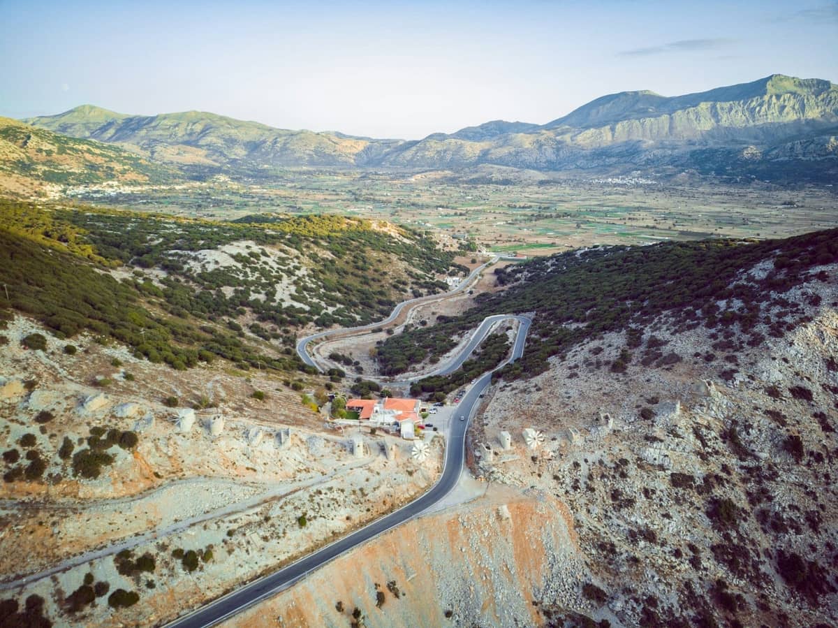 Jalan pegunungan yang berliku dengan pemandangan luas melintasi Dataran Tinggi Lassithi dan perbukitan sekitarnya di Kreta, ideal untuk perjalanan pemandangan alam dan tur multi-pemberhentian.