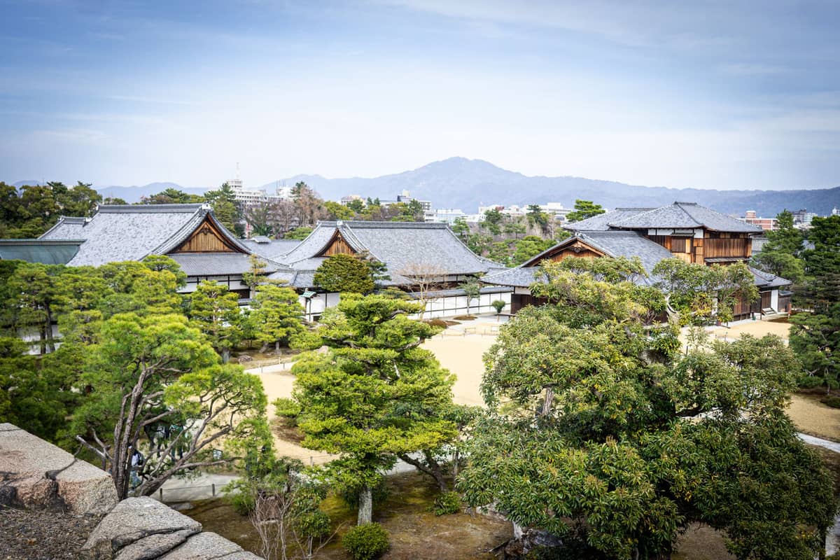 Panoramic view of Nijo Castle’s historic wooden villas surrounded by landscaped stroll gardens and manicured trees in central Kyoto, with mountains in the background.