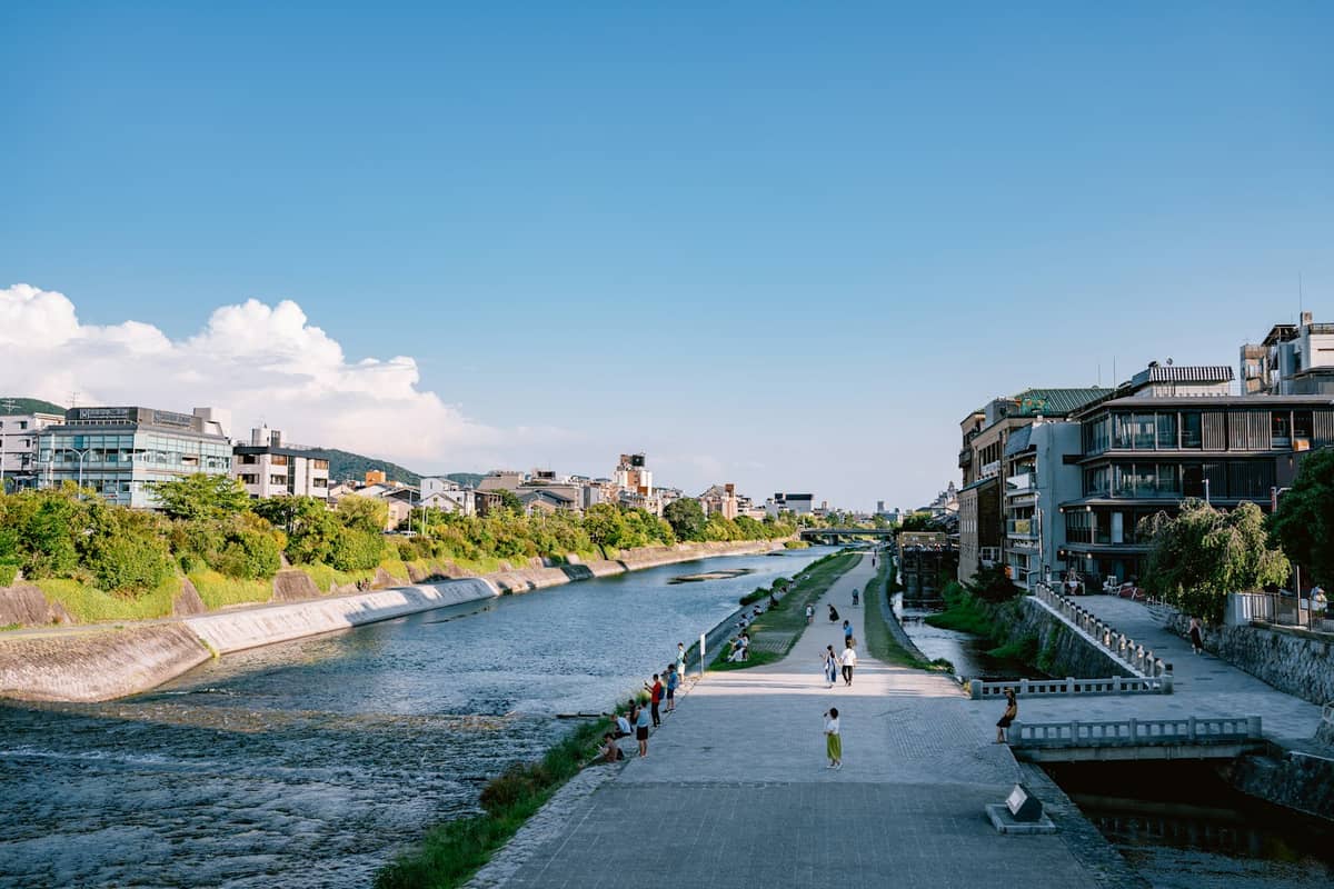 Wide view of Kamo River in Kyoto bordered by green spaces and traditional and modern buildings, with people walking and relaxing along the riverside paths.