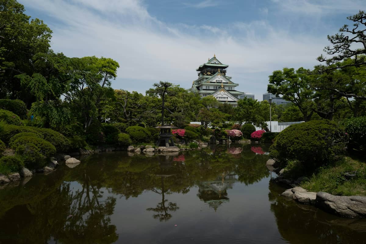 Osaka Castle seen across a landscaped pond with lush greenery and manicured trees in the foreground.