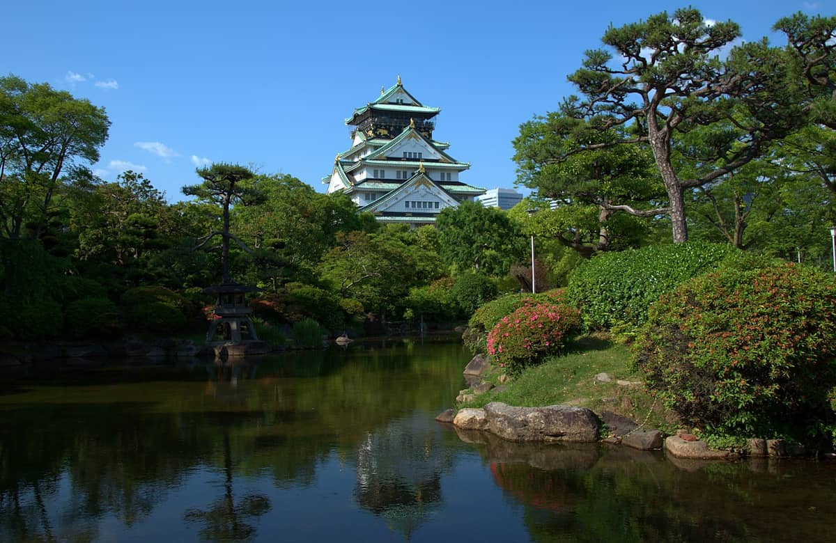 Osaka Castle surrounded by lush greenery and a traditional Japanese garden with a pond reflecting the castle and trees.