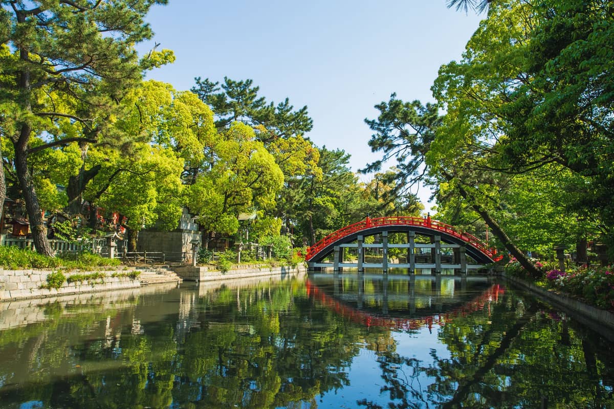 A vivid red arched bridge over a reflective pond surrounded by lush greenery at a shrine garden in Osaka.