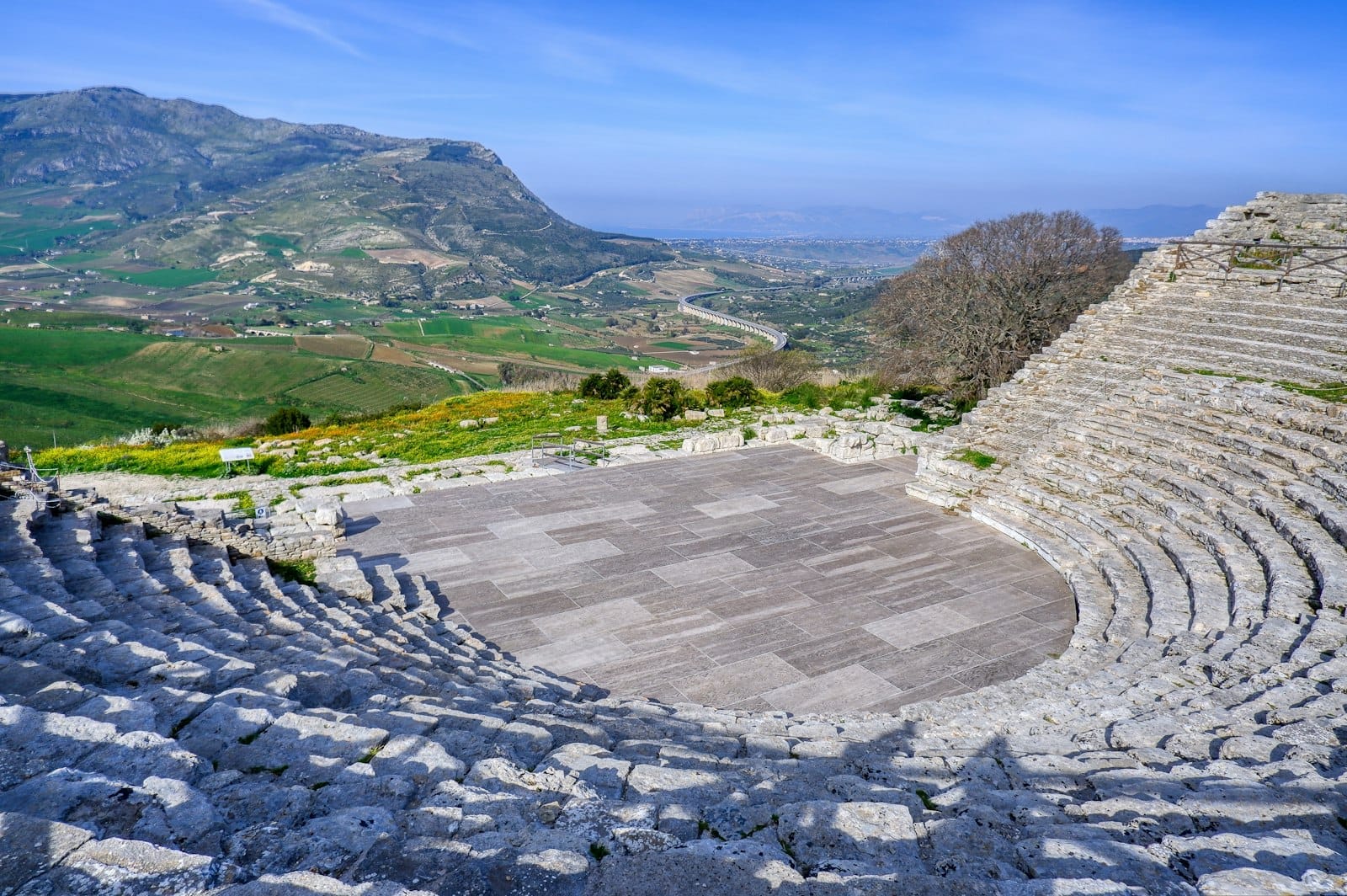 Weitwinkelansicht der Ruinen eines antiken griechischen Theaters in Sizilien, umgeben von malerischen grünen Hügeln und fernen Tälern unter einem klaren Himmel.