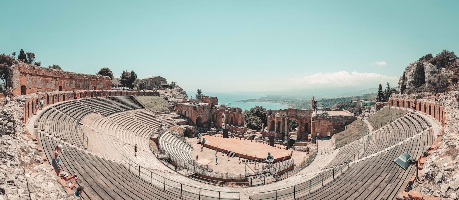 Weites Panorama des antiken griechischen Theaters in Taormina, Sizilien, mit steinernen Sitzreihen, Bühnenruinen und der im Hintergrund sichtbaren Küstenlinie.