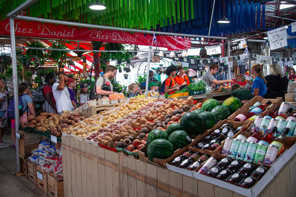 Interno affollato del mercato alimentare di Montreal con prodotti freschi, acquirenti e venditori sotto colorate decorazioni appese.