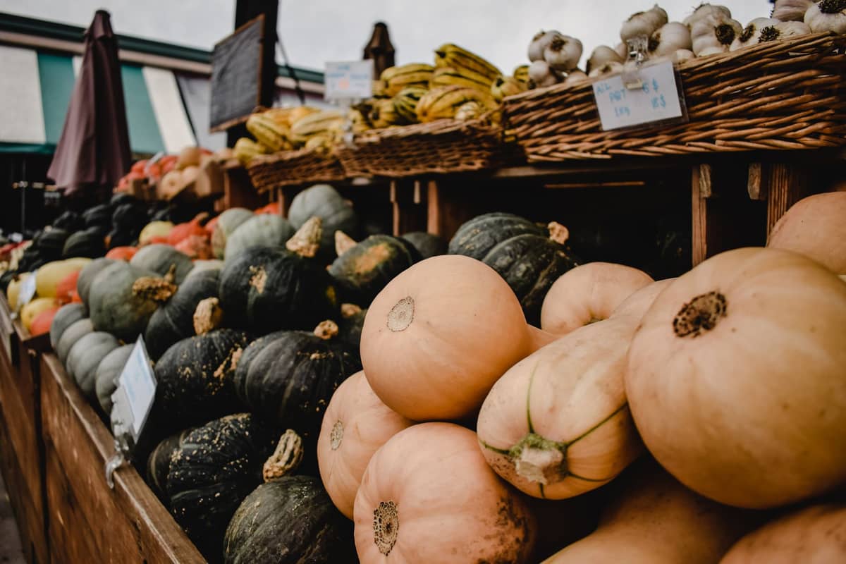 Bancarella di mercato all'aperto con mucchi di zucchine, zucche, banane e aglio in cesti presso un mercato ortofrutticolo.