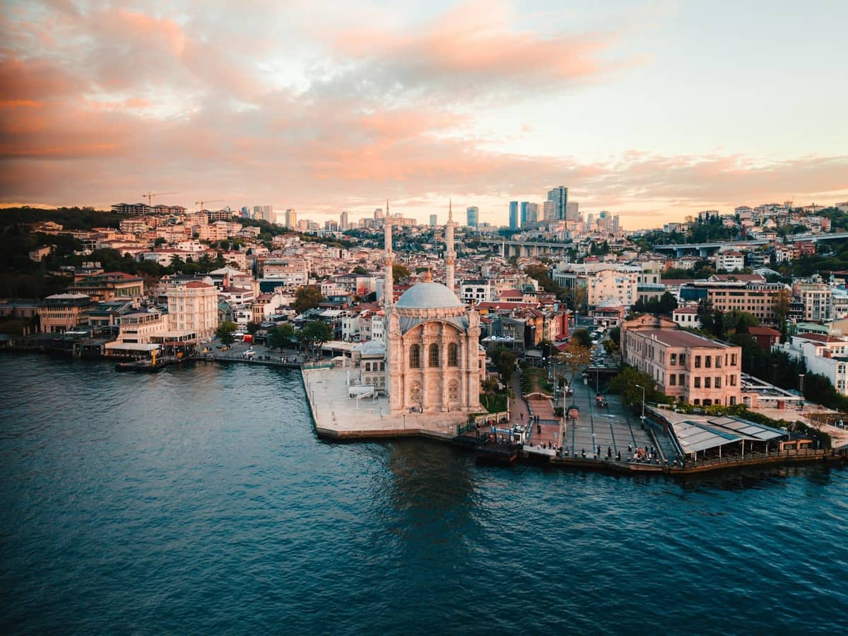 Vista aérea de una mezquita frente al mar en Estambul, rodeada de edificios de la ciudad y el Bósforo, con un puente al fondo al atardecer.