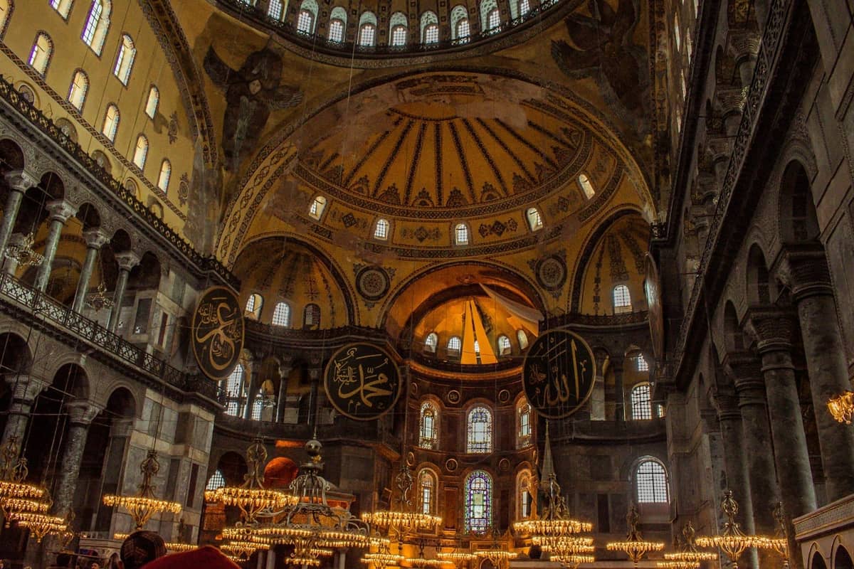 Vista amplia de la gran cúpula y las galerías superiores de Hagia Sophia, con grandes medallones caligráficos y detalles arquitectónicos históricos dorados.
