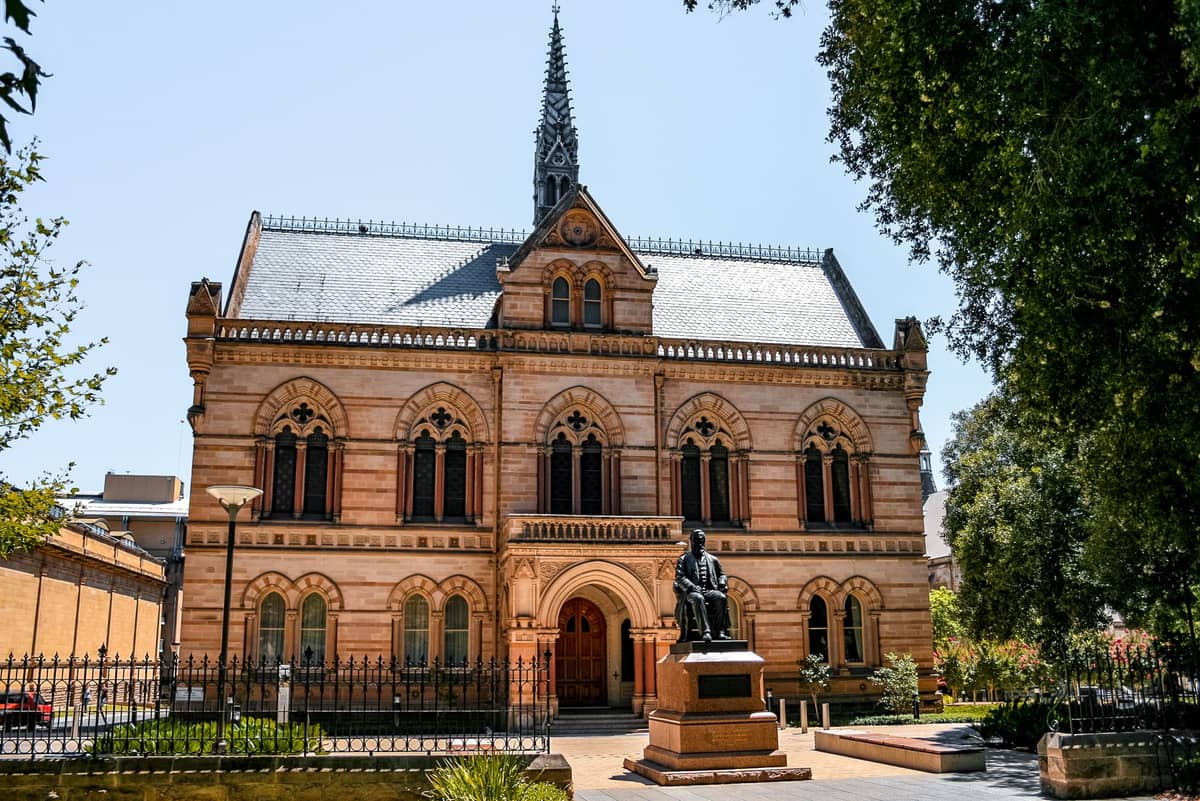 Historisches Sandsteingebäude mit gotischen Fenstern und einem Turm, Statue davor, Bäume rahmen die Szene—Elder Hall an der North Terrace in Adelaide.