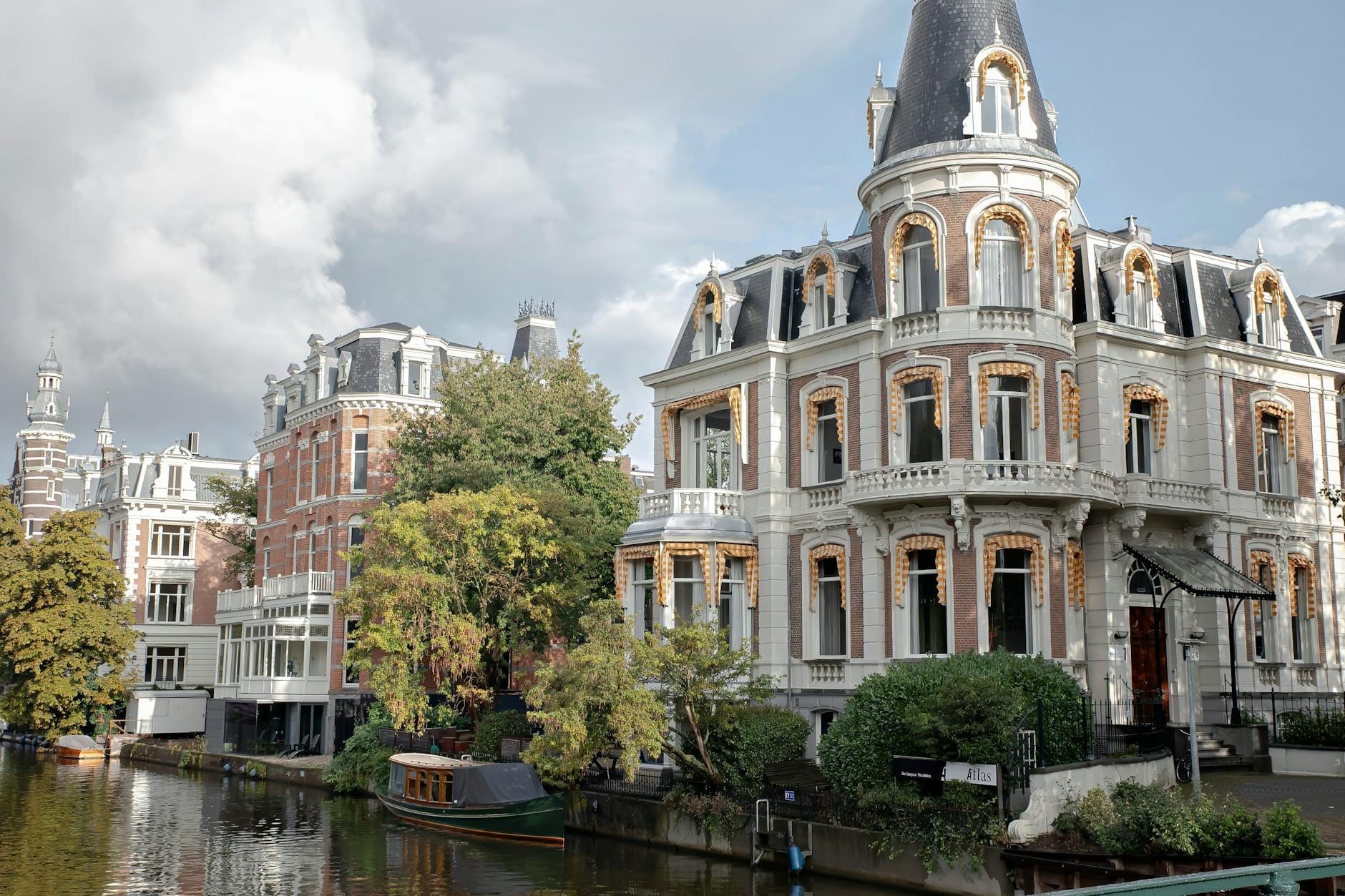 Mansión histórica ornamentada junto a un canal en Ámsterdam, con abundante vegetación, arquitectura clásica y barcas en el canal, bajo un cielo parcialmente nublado.