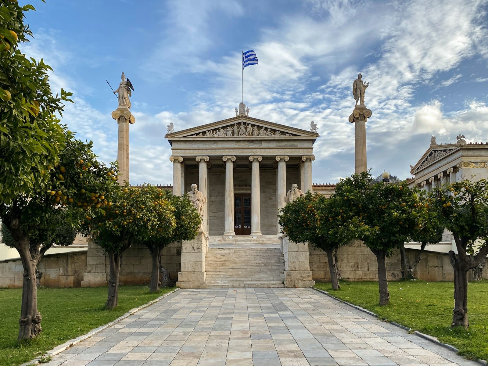 Fachada neoclássica imponente do Museu Arqueológico Nacional de Atenas, com fileiras de colunas e estátuas sob um céu azul e a bandeira grega ao vento.