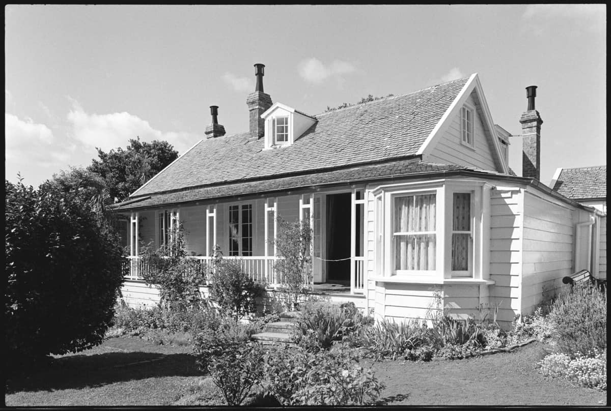 Villa de madeira histórica com varanda e jardim de flores, típica da arquitetura patrimonial vista no Albertland Heritage Museum ou em locais similares em Auckland.