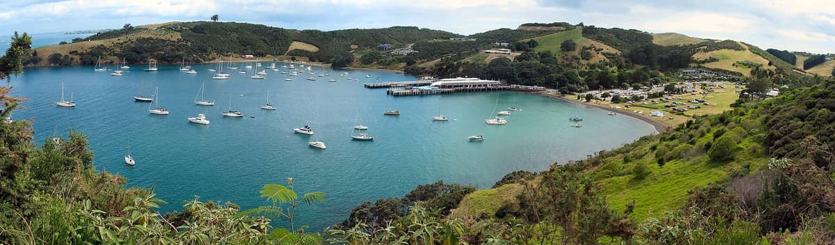 Vista panorâmica da ilha Waiheke com um cais movimentado, barcos atracados numa baía turquesa e colinas verdes ao fundo.