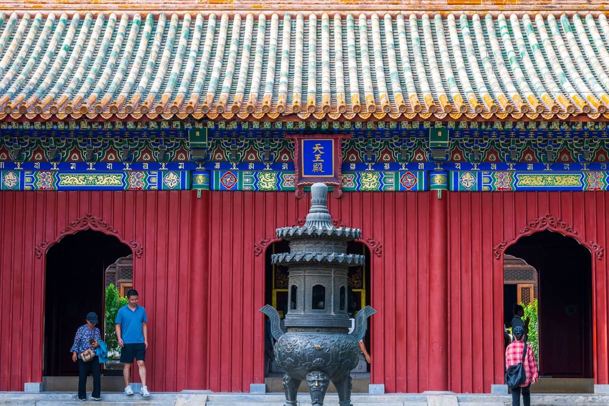 Außenansicht eines traditionellen chinesischen Tempels in Beijing mit einem bronzenen Räuchergefäß und Menschen, die an dem prachtvoll dekorierten roten Gebäude vorbeigehen.