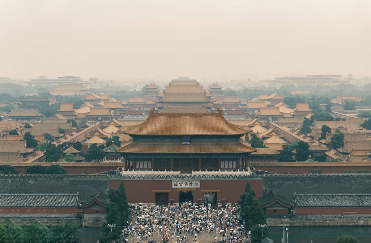 Weitläufige Draufsicht auf den Palastkomplex der Verbotenen Stadt in Beijing, mit goldenen Dächern, Toren und Menschenmassen am Eingang unter einem dunstigen Himmel.