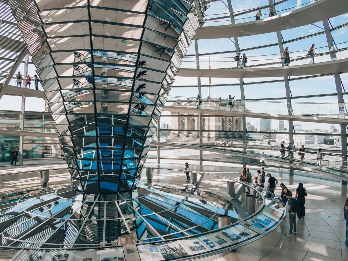 Weitwinkelansicht des Innenraums mit Glaskuppel des Berliner Reichstags, mit Besuchern, die an geschwungenen Rampen entlanggehen, und einfallendem Tageslicht.