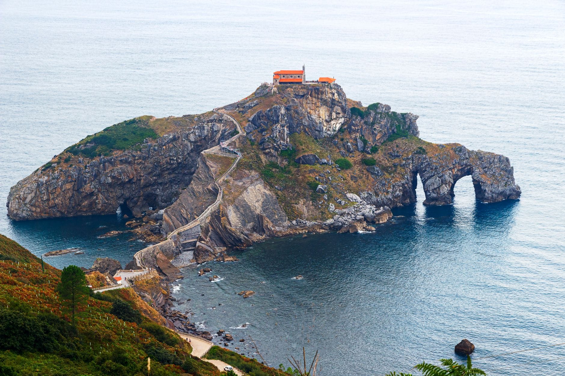 Dramatyczny widok na wysepkę Gaztelugatxe z krętą kamienną ścieżką i kaplicą, otoczoną błękitnym morzem i skalistym wybrzeżem.