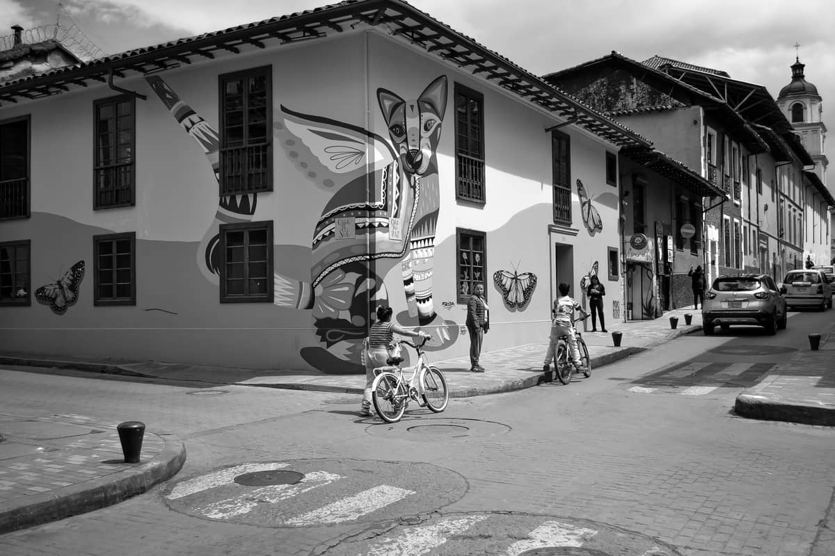 Black and white photo of a mural-covered building on a street corner in Bogotá, with cyclists and pedestrians passing by.