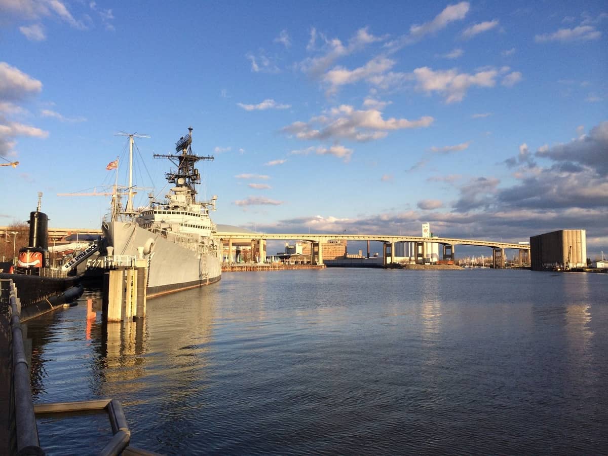 Destructor naval desafectado atracado en el frente marítimo de Boston bajo un cielo parcialmente nublado, con edificios y puentes del paseo marítimo al fondo.