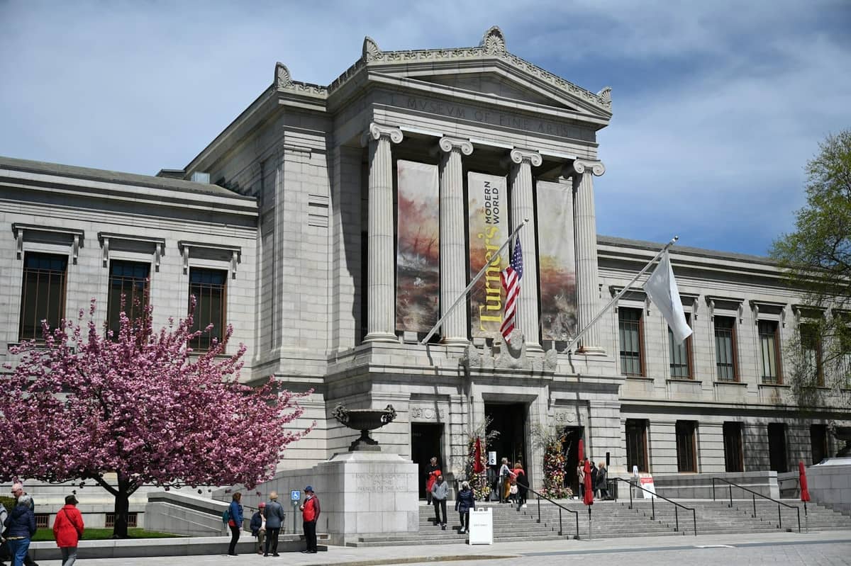 Vista frontal del Museum of Fine Arts Boston con columnas, pancartas, árbol en flor y personas afuera en un día soleado.