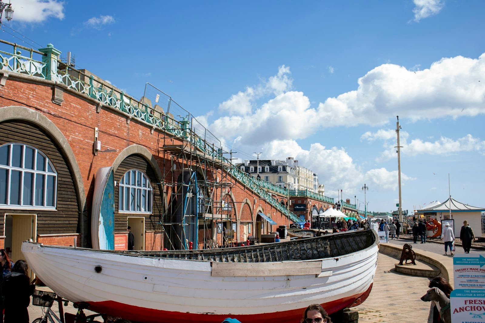 View of Brighton seafront with a historic brick arcade, blue sky, and a traditional fishing boat, evoking Brighton’s maritime and heritage scene.