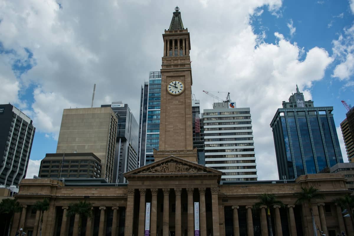 Brisbane City Hall na may iconic na clock tower, mga klasikong haligi, at mga skyscraper sa likod sa isang maliwanag na araw.