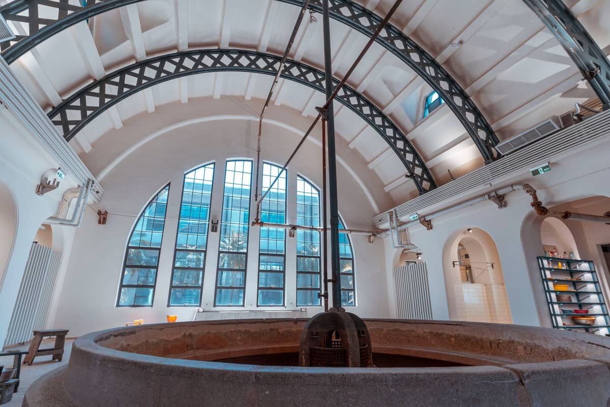 Interior de uma cervejaria histórica ou espaço de museu industrial com grandes janelas arqueadas e teto abobadado, com equipamentos de fabricação de cerveja.